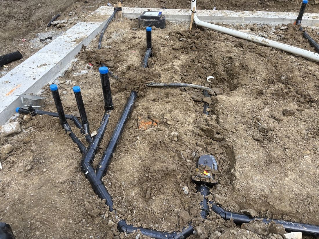 Black plumbing pipes and vertical stub-ups laid in the ground beside a concrete foundation wall